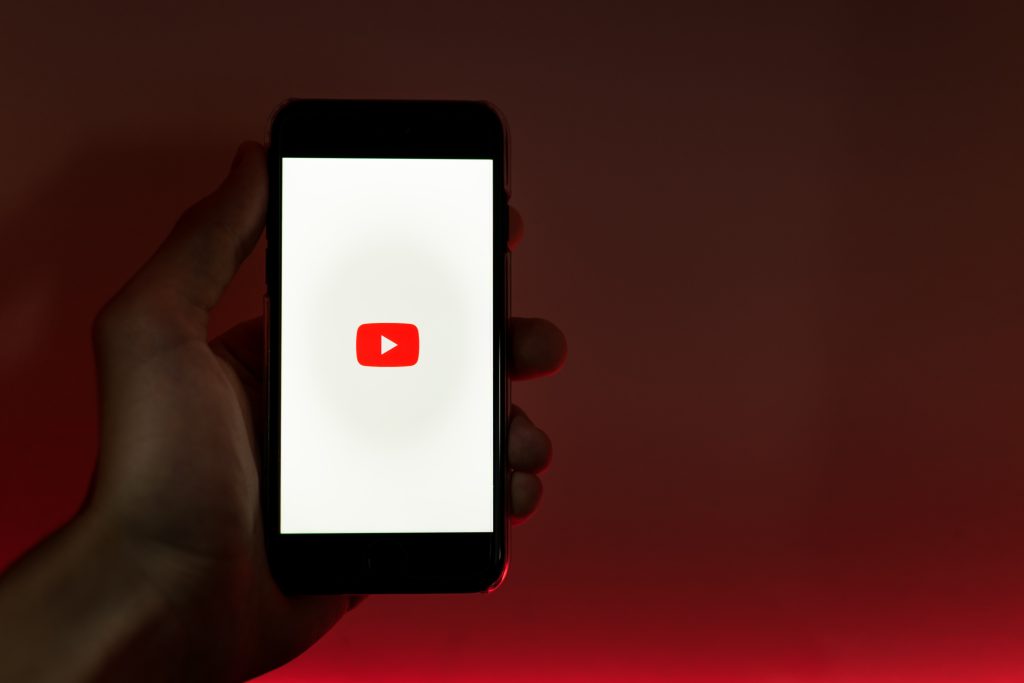 Mão segurando um celular com a tela ligada na página inicial do Youtube. Fundo da foto vermelho. 